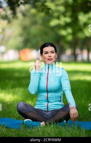 Foto verticale. Ritratto esterno a lunghezza intera di una donna in posizione lotus inserisce gli auricolari per ascoltare la musica per il relax. Foto Stock