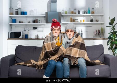 coppia fredda, avvolta in coperta di plaid, indossando cappelli caldi, guardando la macchina fotografica mentre si siede sul divano in cucina Foto Stock