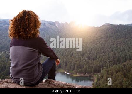 Escursionista maschile con capelli ricci, seduto sul bordo della scogliera, ammirando l'alba Foto Stock