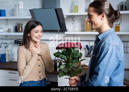 uomo che presenta le rose alla donna soddisfatta che tiene le mani vicino al petto mentre in piedi in cucina Foto Stock