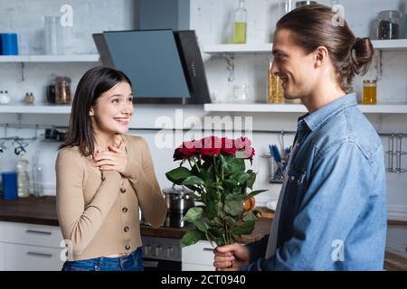 giovane uomo che presenta bouquet di rose a donna felice tenuta mani vicino al petto in cucina Foto Stock