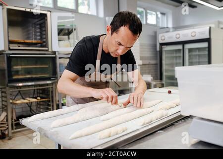 Acuto panettiere maschio che fa baguette su una tavola fiorita dentro una cucina Foto Stock