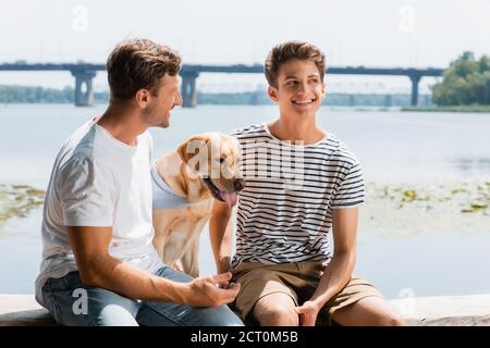 padre che guarda un figlio compiaciuto vicino al golden retriever e al fiume Foto Stock