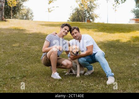 padre abbracciando ragazzo adolescente vicino golden retriever mentre si siede erba nel parco Foto Stock