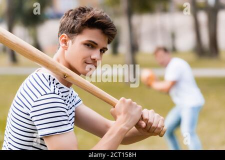 fuoco selettivo del figlio adolescente con il bat di softball che guarda macchina fotografica mentre si gioca a baseball con il padre nel parco Foto Stock