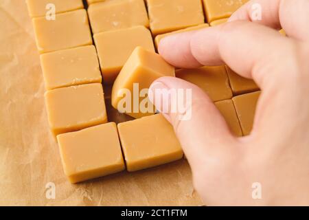 La mano della donna prende un caramello di vaniglia da una carta marrone pergamena. Caramelle fresche e gustose a base di latte e zucchero. Pezzi quadrati di delizioso fudge. Foto Stock