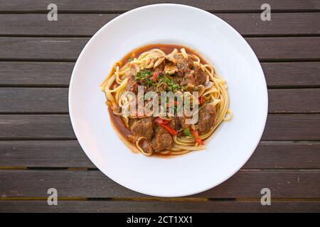 Ragù di manzo con peperoni rossi e funghi con contorno di prezzemolo su pasta fatta in casa in un piatto bianco su un rustico tavolo di legno scuro, vista ad angolo alto Foto Stock