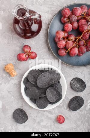 Patatine nere con carbone, aceto balsamico in vetro, uva rossa su un piatto di ceramica blu su fondo grigio di cemento. Vista dall'alto, disposizione piatta, clo Foto Stock