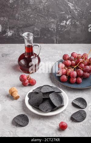 Patatine nere con carbone, aceto balsamico in vetro, uva rossa su un piatto di ceramica blu su fondo grigio di cemento. Vista laterale, spazio copia, Foto Stock