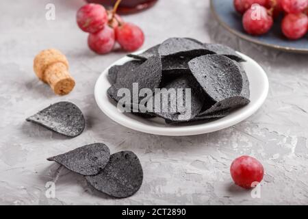 Patatine nere con carbone, aceto balsamico in vetro, uva rossa su un piatto di ceramica blu su fondo grigio. Vista laterale, primo piano, se Foto Stock