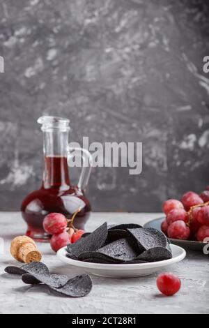 Patatine nere con carbone, aceto balsamico in vetro, uva rossa su un piatto di ceramica blu su fondo grigio. Vista laterale, primo piano, se Foto Stock