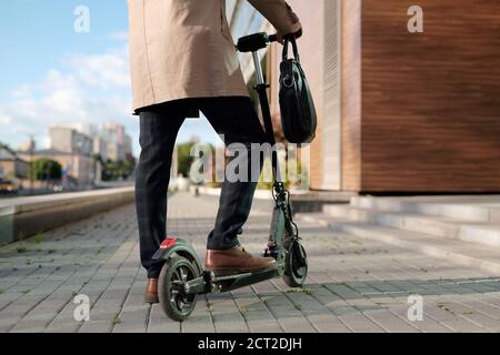 Sezione bassa di uomo d'affari in trenchcoat beige e pantaloni in piedi su scooter Foto Stock