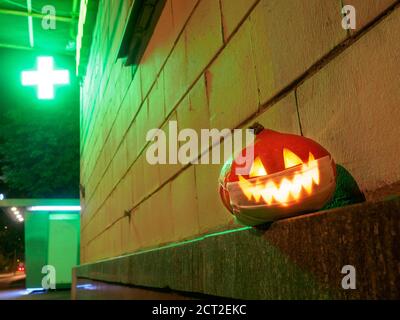 La zucca di Halloween con una maschera protettiva sul viso sullo sfondo di una croce farmacia che si illumina al buio. Concetto di Halloween durante il Foto Stock