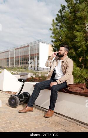 Giovane uomo indolente in elegante casualwear che parla su smartphone all'aperto Foto Stock