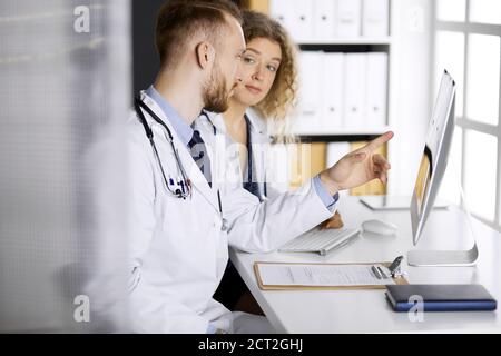 Medico arrossato con collega femminile che parla della terapia attuale della malattia mentre si siede al posto di lavoro in clinica. Lavoro di squadra in medicina Foto Stock