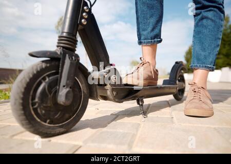 Gambe di giovane donna in scarpe beige e jeans blu in piedi su uno scooter elettrico Foto Stock