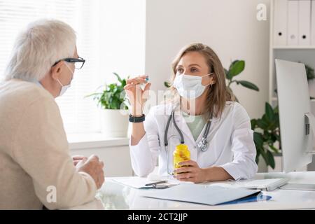 Giovane medico fiducioso in whitecoat e maschera protettiva mostrando pillola al paziente Foto Stock