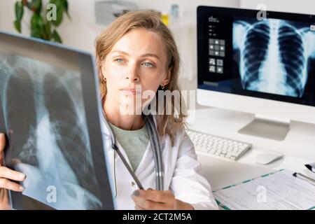 Radiologo femminile giovane e serio nel whitecoat che punta all'immagine radiografica di polmoni Foto Stock