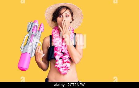 Bella giovane donna con capelli corti indossando bikini e hawaian lei tenendo pistola acqua coprendo la bocca con la mano, scioccata e impaurita per errore. Surp Foto Stock