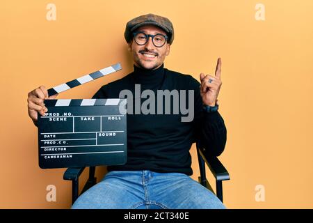 Bell'uomo con tatuaggi che tengono la clapboard del video film seduto sopra la sedia del direttore sorride felice che punta con la mano ed il dito a. il lato Foto Stock