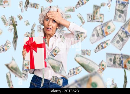 Donna anziana dai capelli grigi che tiene il regalo stressato e frustrato con la mano sulla testa, sorpresa e faccia arrabbiata Foto Stock