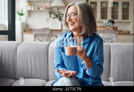 Felice rilassata donna anziana matura bere caffè rilassante sul divano a casa. Foto Stock