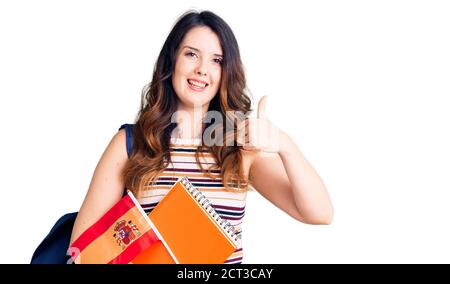 Bella giovane donna brunette scambio studente tenendo bandiera spagna sorridente felice e positivo, pollice su facendo eccellente e segno di approvazione Foto Stock