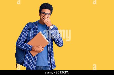 Giovane afroamericano uomo che indossa zaino studente tenendo libro ridendo e imbarazzato ridere bocca copertura con mani, pettegolezzi e concetto di scandalo Foto Stock