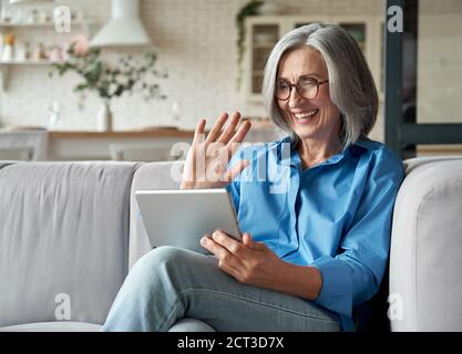 Happy anni '60 donna matura che agita la mano tenendo le videochiamate digitali da tablet a casa. Foto Stock