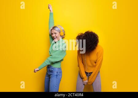 Donne carine con cuffie che ballano su una parete gialla dello studio pieno di emozioni con pullover caldi e jeans Foto Stock