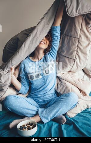 Bella donna con capelli ricci si sta stretching in letto coperto di una trapunta prima di mangiare i cereali Foto Stock