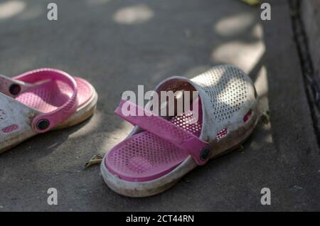 Pantofole estive sporche di colore bianco-rosa all'ombra su un percorso in cemento. Primo piano delle vecchie pantofole. Messa a fuoco selettiva Foto Stock