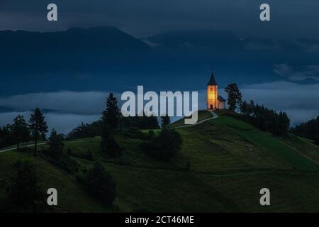Jamnik, Slovenia - ora blu a Jamnik con la chiesa illuminata di San Primoz in cima a una nebbia alba estiva. Alpi Giulie sullo sfondo Foto Stock