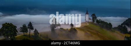 Jamnik, Slovenia - Foggy alba estiva nella chiesa di Jamnik St.Primoz. La nebbia passa dolcemente dalla piccola cappella in cima ad una vasta fotografia panoramica Foto Stock