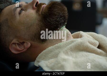 Uomo bearded che ottiene il taglio dei capelli dal parrucchiere al barbershop. Affilato. Crema per la rasatura in legno di sandalo. Balm su parti del corpo asciutte per idratare la pelle. Vecchio Foto Stock