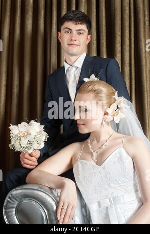 Sposa con fiore in capelli e sposo seduto dietro tende marroni ripiegate Foto Stock