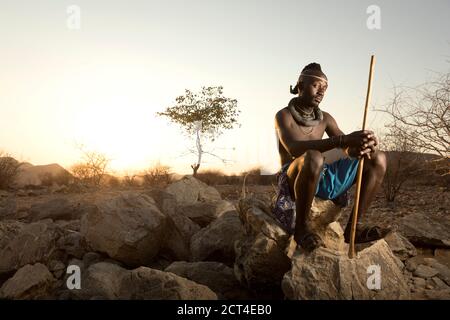Un uomo Himba si siede su una roccia mentre il sole tramonta alla fine della giornata a Epupa, nella regione di Kunene, in Namibia. Foto Stock