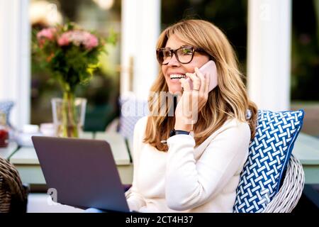 Ritratto di felice donna matura con un notebook e parlando con qualcuno sul suo telefono cellulare mentre si siede sul balcone a casa. Foto Stock