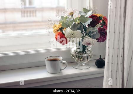 La natura morta dell'autunno femminile. Tazza di caffè, bouquet di fiori colorati di dahlia in vaso di vetro sul davanzale. Composizione floreale moody con fico fr Foto Stock