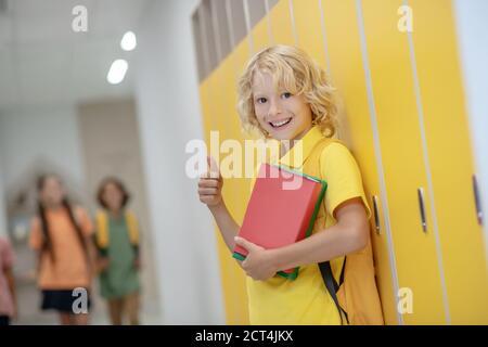 Ragazzo biondo sorridente in camicia gialla in piedi vicino agli armadietti nel corridoio Foto Stock