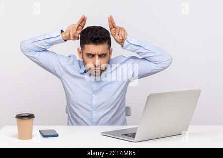 Conflitto sul lavoro. Uomo aggressivo dipendente seduto in ufficio e mostrando bull Horn gesto, minacciando di attaccare, guardando anglicamente alla fotocamera. In Foto Stock