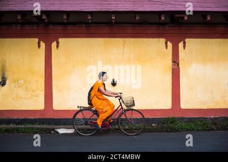 Scena di strada indiana di vita locale fuori di un tempio con una donna in bicicletta con una parete gialla, Fort Kochi (Cochin), Kerala, India Foto Stock