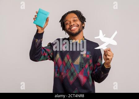 Allegro ragazzo africano sorridente con dreadlock che tiene in mano passaporto e aereo di carta, sognando di vacanza all'estero, avventure. Riprese in studio al coperto Foto Stock