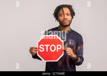 Interessato africano ragazzo con Dreadlock che tiene rosso stop segno in mani, attenzione, sia concetto attento, attenzione. Studio al coperto isolato sulla parte posteriore grigia Foto Stock