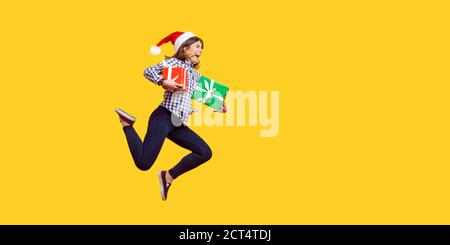 Vista laterale ritratto di estremamente gioiosa brunette donna in santa hat jumping o in esecuzione in aria con avvolto xmas confezioni regalo, celebrando sconti. indo Foto Stock