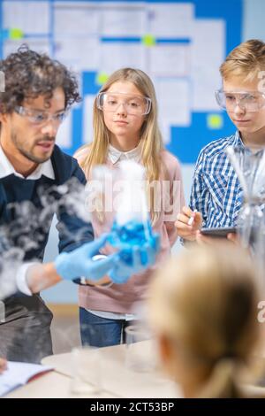 Scuola elementare Scienza Aula: Insegnante entusiasta spiega la chimica a diversi gruppi di bambini, mostra loro come mescolare le sostanze chimiche in Beakers Foto Stock