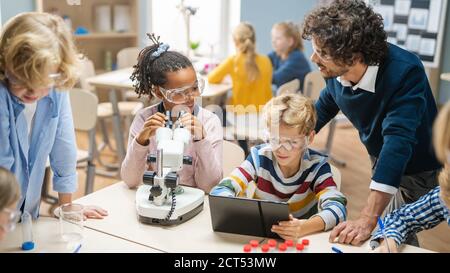 Scuola elementare Scienza Classroom: Cute Little Girl guarda sotto Microscopio, Boy usa Digital Tablet computer per controllare le informazioni su Internet Foto Stock