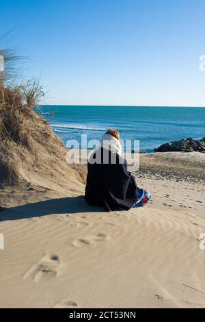 vista posteriore della donna vestita in abito invernale seduto dune di sabbia su una spiaggia che si affaccia sul mediterraneo mare in una giornata di sole Foto Stock