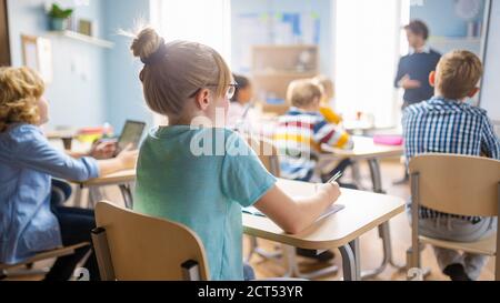 Classe di scienza della scuola elementare: Cute Little Girl sta ascoltando un insegnante e facendo note. L'insegnante di fisica spiega la lezione a una classe diversa piena Foto Stock