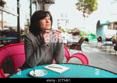 Donna matura di affari siedono da sola e pensando. Tenendo una tazza bianca di tè o caffè, guardare a lato. Pensare durante una breve pausa. Penna con notebook Foto Stock